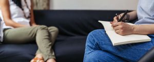 A woman on a sofa speaking to a therapist who is taking notes on a legal pad